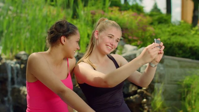 Two girls grimacing when taking photo with phone. Self portrait. Young women posing when taking photo in park. Female friends take photo with smart phone camera. Fitness women taking selfie