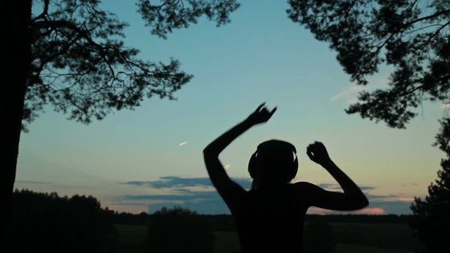 Woman listening to music in headphones, jumping and dancing in the forest after sunset. Freedom and happiness concept.