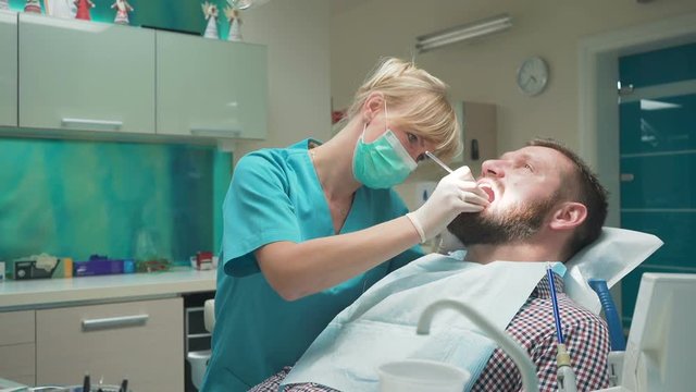 Female dentist examining an drilling patient's tooth. Visit is in proffessional dental clinic. He is sitting on dental chair. He is young and has beard. Dolly and steadicam shot.
