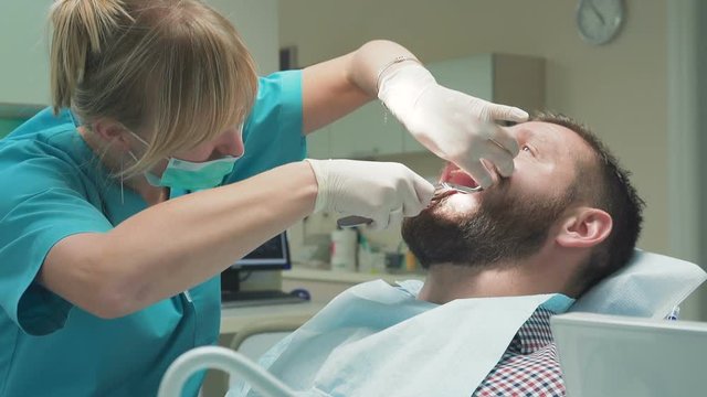 Female dentist examining an drilling patient's tooth. Visit is in proffessional dental clinic. He is sitting on dental chair. He is young and has beard. Dolly and steadicam shot.
