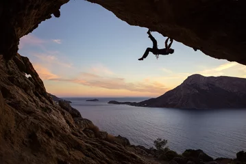 Gordijnen Male climber climbing along roof in cave against beautiful view of coast below  © Andrey Bandurenko