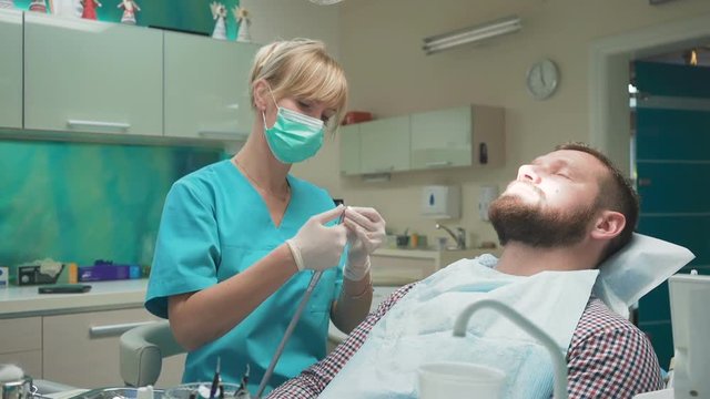 Female dentist examining an drilling patient's tooth. Visit is in proffessional dental clinic. He is sitting on dental chair. He is young and has beard. Dolly and steadicam shot.
