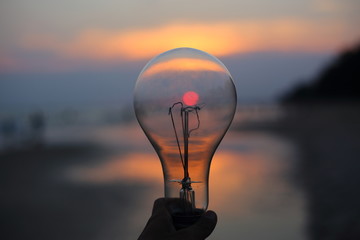 hand holding a electric light bulb and sun at beach sunset sky
