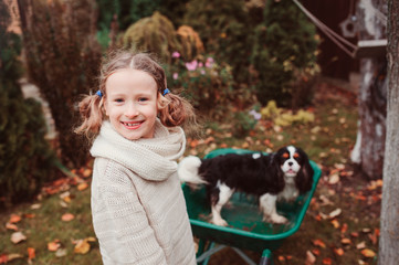 Naklejka na ściany i meble happy funny child girl riding her dog in wheelbarrow in autumn garden, candid outdoor capture, kids playing with pets