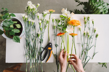 Florist workspace: caucasian woman making floral decorations. Top view of flowers and greenery on...