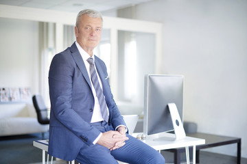 Shot of a senior investment advisor businessman sitting on desk at office and looking at camera.