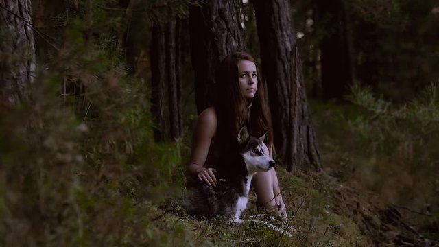 Enigmatic young woman sitting in the forest with beautiful dog and looking at the camera