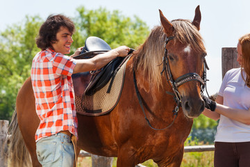 Obraz premium Young man and woman saddling chestnut brown horse