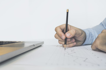 Close-up of a black pencil in a man's hand. Businessman takes notes in pencil on the schedule. On a white desktop is a laptop. The image on white background, isolated. On the table is digital gadget.