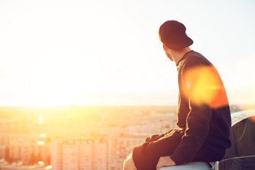 Young and brave man sitting on the edge of the roof and looking far away at the city, lens flares