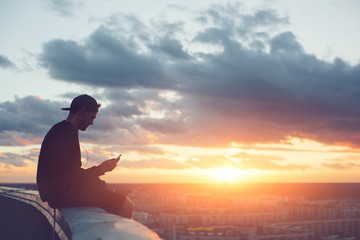 Risky man with smartphone chilling on the edge of the roof at sunset
