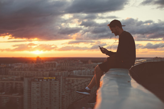 Young brave man sitting on the edge of the roof with smartphone
