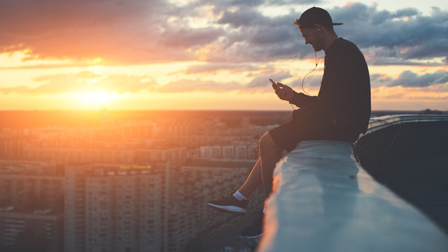 Young brave man sitting on the edge of the roof with smartphone at sunset