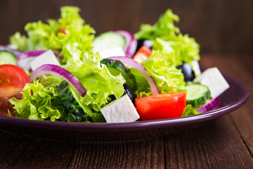 Greek salad (lettuce, tomatoes, feta cheese, cucumbers, black olives, purple onion) on dark wooden background close up. Healthy food.