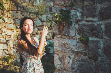 Feminine portrait of young romantic woman with long hair, red lips and manicure in white dress flowers. Pretty female in Stari Bar old fortress, Montenegro. Brunette girl walk around castle