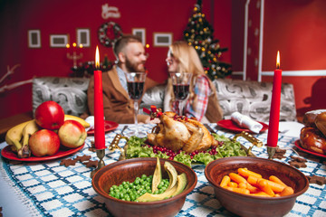 Beautiful couple in a decorated festive interior with a Christmas tree drinking wine. A romantic dinner for thanksgiving with fried chicken and candles