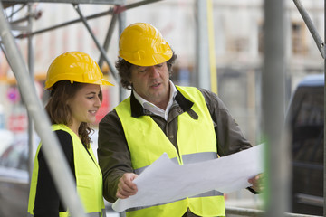 Male and female architects checking blueprints among scaffolding on construction site