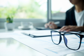 Office desk table with eyeglasses focus and asian business woman working background. Vintage tone Retro filter effect,soft focus,low light.(selective focus).
