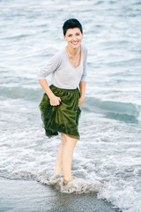 Portrait of beautiful smiling laughing Caucasian brunette woman with short hair in grey shirt, green olive tutu tulle skirt, standing barefoot on beach in sea water, free happy lifestyle