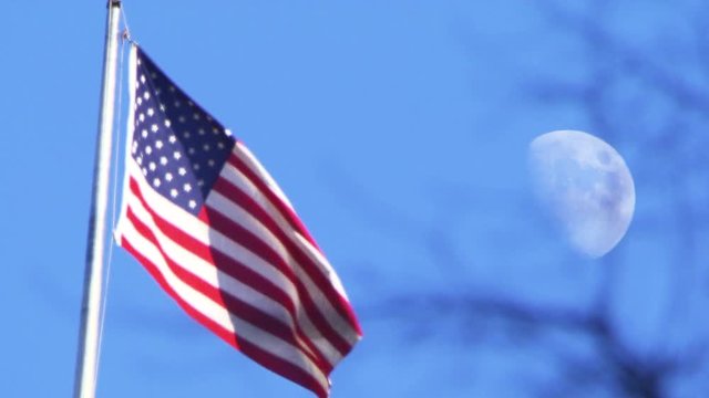 American flag waves in wind, moon in background, pull focus day time shot.