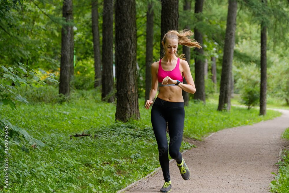 Wall mural Young slim woman jogging in a park looking to smart watch