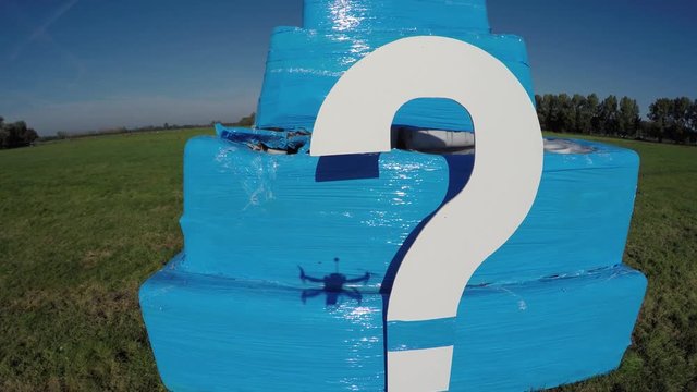 Aerial of white question mark on small bright blue structure drone hoovers then flying backwards away from cube with ?  also showing quadcopter shadow footage could indicate FAA drone regulations 4k