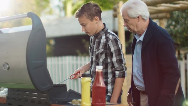 Grandfather and Grandson Are Cooking Burgers on Grill at Bright Summer Day.