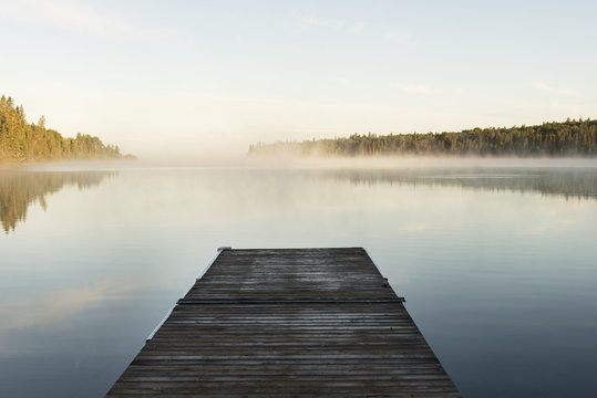 A wooden dock leading out to a misty tranquil lake in Riding Mountain National Park; Wasagaming, Manitoba, Canada