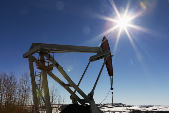 Pumpjack and snow covered hills with sun burst and blue sky; Longview, Alberta, Canada