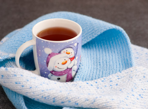 Кружка чая с изображением снеговика и снега в окружении теплого шарфа. Чай,  вязаный шарф, рисунок снеговика и снега напоминают о приятных ощущениях, когда можно согреться во время холода.