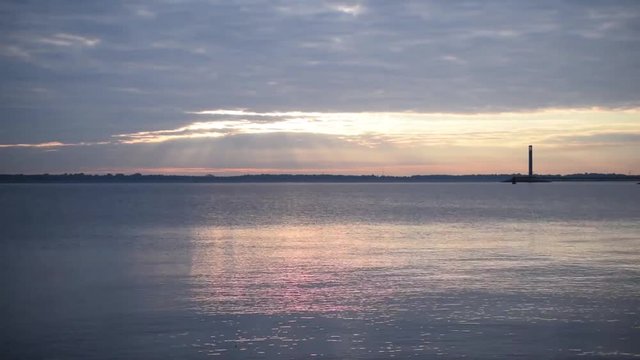 Time lapse of colorful clouds moving over water at dawn with lighthouse and beautiful light coming through clouds