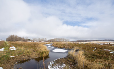 A stream dotted with snow patches winding through a harvested field under cloudy sky in rural landscape