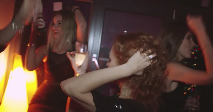 Shaky cam of tipsy beautiful young women with clubbing makeup on holding alcohol and dancing suggestively alongside with men in stylish suits 