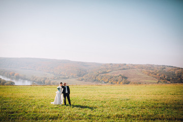 Bride and groom walking and holding hands on a meadow. Wide angle sunset photo. Autumn pcture. River and mountain landscapes