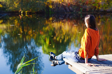 Beautiful young woman sitting on the edge of wooden jetty by the lake with autumn reflections