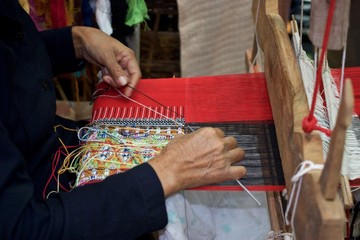 Old woman weaving red black and multi color cotton flag pattern on loom in North of Thailand