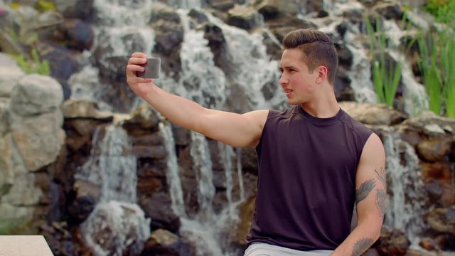 Gay taking selfie near waterfall. Man posing for selfie photo outdoor. Teenager taking photo with phone. Selfie man. Handsome man taking selfie in park. Gay making self portrait. Young man smiling