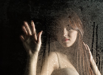 Smooth portrait of sexy model, posing behind transparent glass covered by water drops.