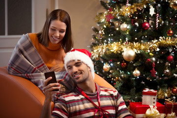 Happy young couple is using smart phone while sitting in front of the Christmas tree at home.