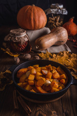 Autumn composition with roast pumpkin and garlic in a cast iron skillet on a wooden table with maple leaves, dried mushrooms, rosehips and whole pumpkins. Photos in rustic style.
