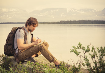 Young Man Traveler with backpack reading book and writing notes outdoor mountains on background Summer vacations and Lifestyle concept
