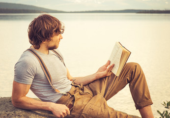 Young Man reading book outdoor with lake on background Summer vacations and Lifestyle concept