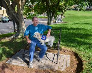 Older gentleman sitting on bench enjoying the sunshine with his Havanese service dog standing on his lap.