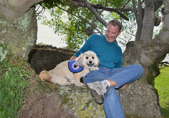 Older gentleman with big smile sitting in huge tree enjoying his Havanese service dog protecting him with tongue sticking out.