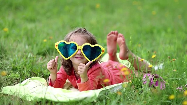Funny little girl in big sunglasses in the shape of hearts lies on green grass. Beautiful little girl in glasses lies on the green grass and looks at the camera