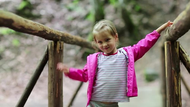 Little girl stands on a wooden bridge and smiles. Funny little girl in pink jacket on the nature