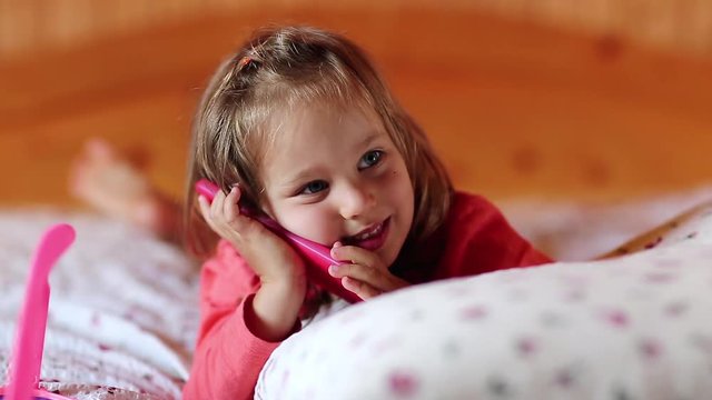 Funny little girl lies on the pillow and holds in hands red mobile phone. Little girl with smartphone lies on the bed and shows his tongue