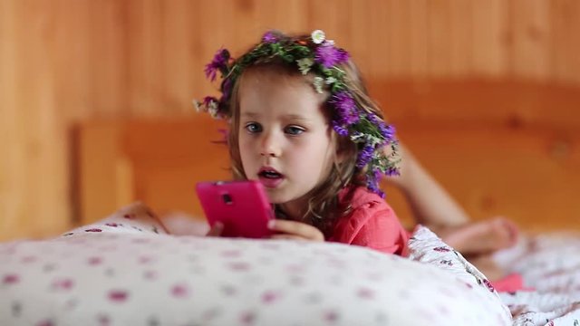 Little girl with a wreath on his head and with red smartphone lies on the bed. Funny little girl lies on the pillow and holds in hands cell phone