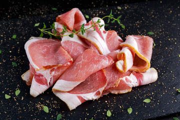 Raw smoked bacon slices on black stone board which herbs