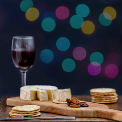 Fototapeta na wymiar Brie and pecan halves on wooden cheese board, accompanied by crackers and one glass of red wine, against black background, decorated with colorful holiday lights- eating alone